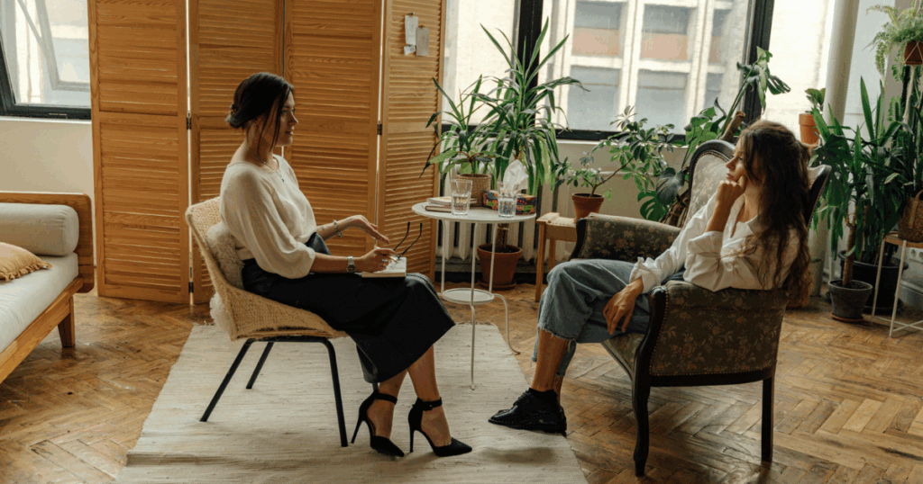 Mental health rehabilitation: woman in therapy session with counselor taking notes, indoor setting, natural light, plants, calm atmosphere.