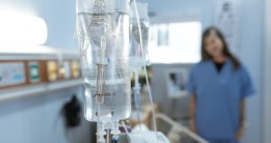 IV drip bags hanging in a hospital room, with a nurse in blue scrubs out of focus in the background.