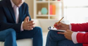 - Los Angeles Mental Health Counselor taking notes on a clipboard while a client sits with hands clasped, in a therapy setting