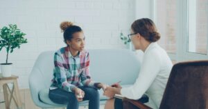 - Los Angeles Mental Health Therapy session: young woman in a plaid shirt talks with a counselor on a light blue sofa, taking notes.