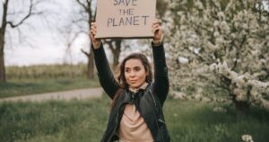 - Los Angeles Mental Health Woman in a black leather jacket raises a cardboard sign reading 'SAVE THE PLANET' in a park with blooming trees behind her.