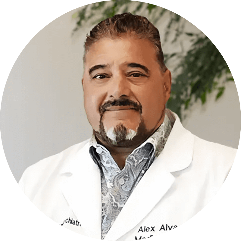 Portrait of Dr. Alex Alva in a white lab coat, smiling, indoors with greenery in the background.