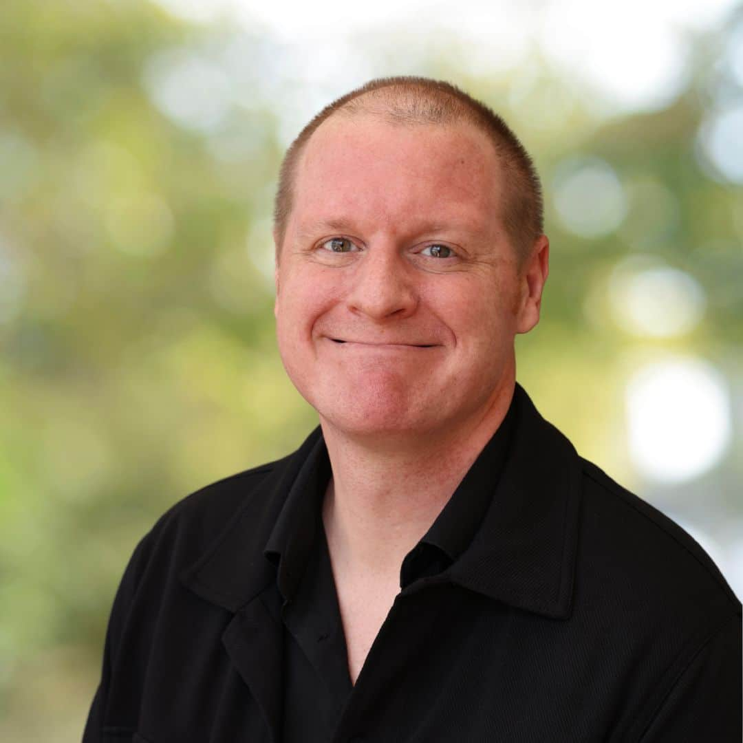 Smiling man with short hair in a black shirt, photographed in a soft-focus outdoor setting.