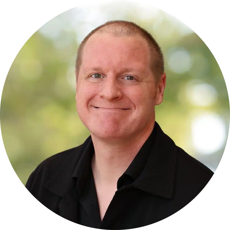 Portrait of a smiling man with short hair wearing a black collared shirt, outdoors.